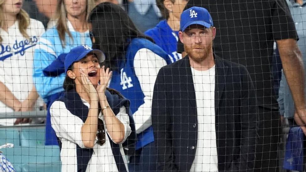 Harry and Meghan standing beside each in the stands of a baseball game, both wearing Dodgers hat, with Meghan cheering
