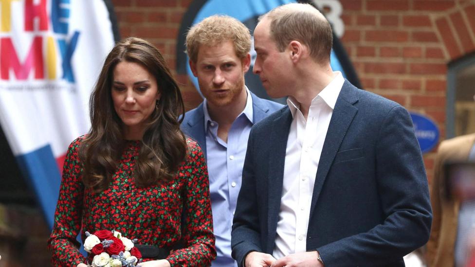 The Princess of Wales, dressed in a red floral dress, and Prince of Wales, in a blue suit, stand in front of the Duke of Sussex