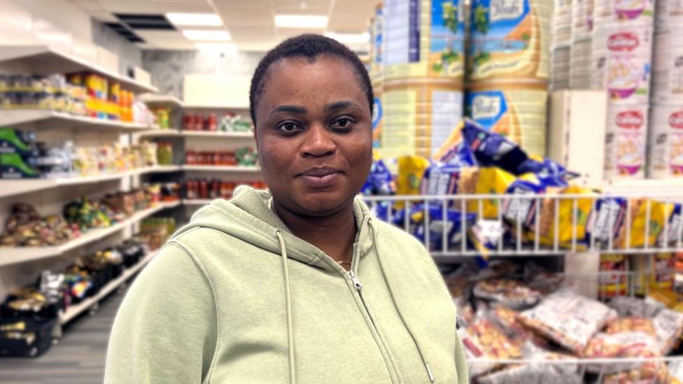 Christianah wears a light green hoodie and stands inside a well‑lit grocery store, with shelves of canned goods, snacks and bulk food items behind her.