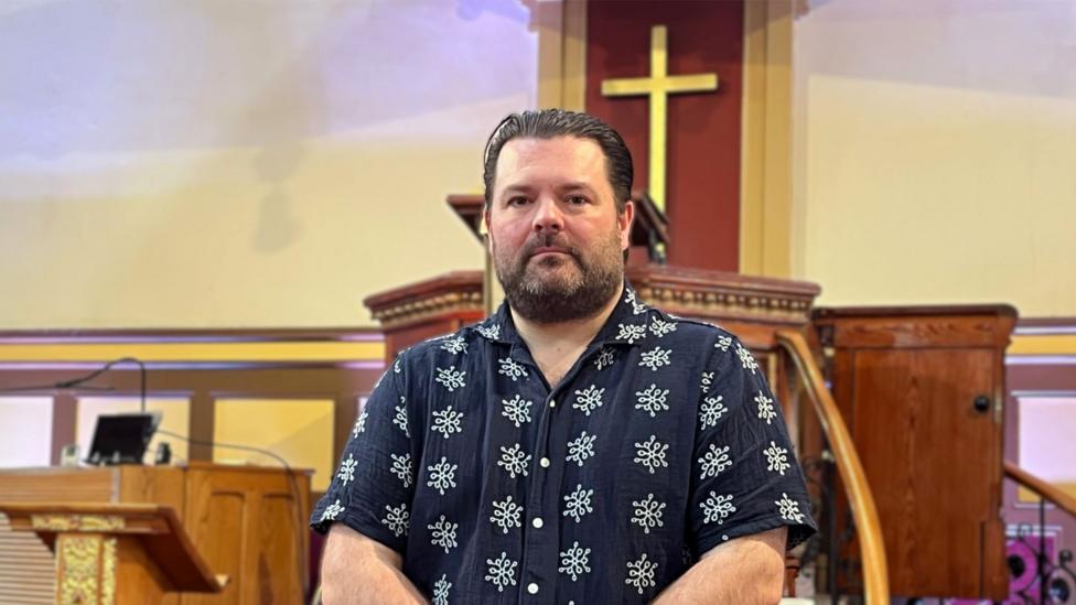 Jonathan stands inside a church in front of a wooden pulpit with a gold cross on a wooden backdrop.