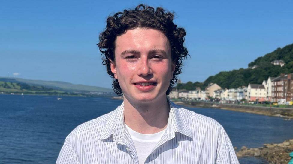 Matthew Quinn stands by a waterfront with calm blue water, coastal buildings and green hills behind him.