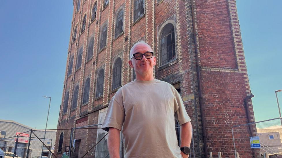Derek Mitchell stands in front of an old brick industrial building with tall arched windows and metal fencing.
