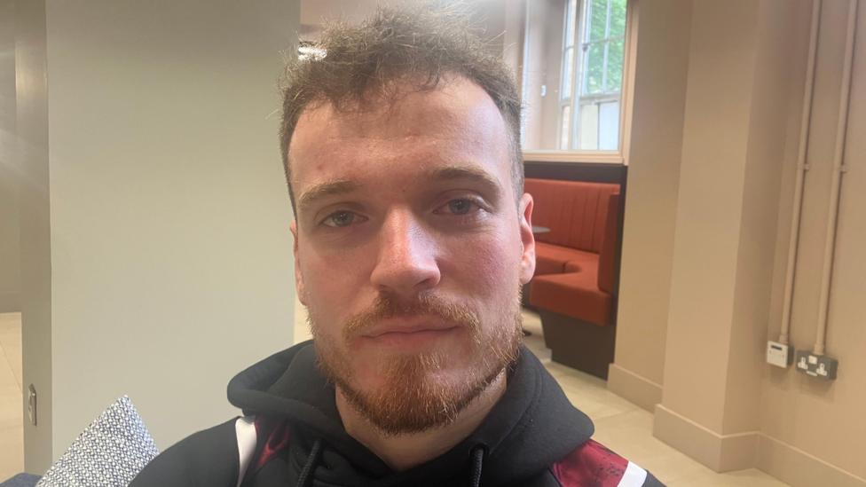 A young man, Caoilte Hamil wearing a black hoodie with red and white on it. He has brunette hair and a brunette beard and moustache.