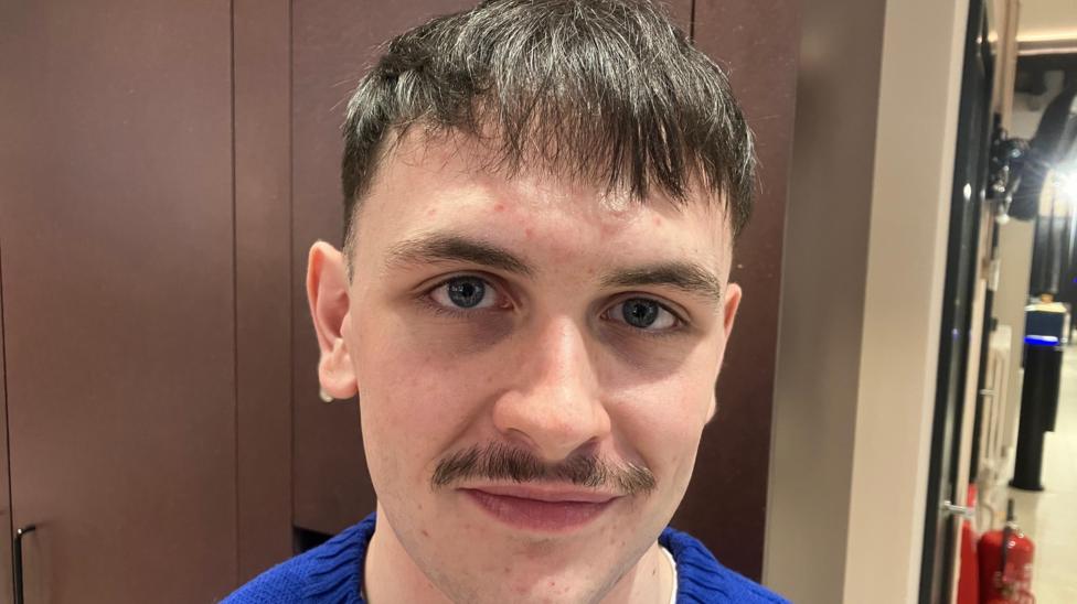 A young man, Turlough O'Hagan pictured wearing a blue jumper. He has brown hair and a dark moustache.