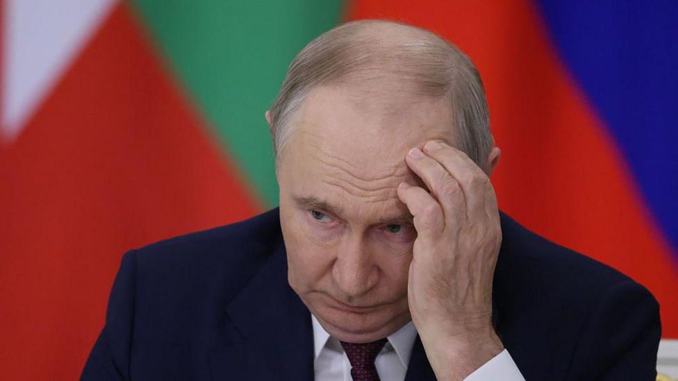 Vladimir Putin sits hunched over in a dark blue suit and red spotted tie, with his left hand to his forehead. He looks tired and nervous.