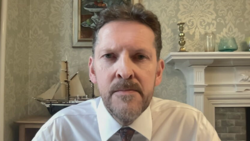 "Dr Ian Kennedy wearing a white shirt and patterned tie, facing the camera in a room with patterned wallpaper, a model sailing ship, framed artwork and a white fireplace mantel in the background."
