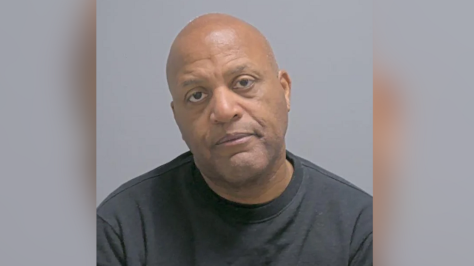 Raymond Williams a man who is sitting down having his picture taken. He is bald and is wearing a black sweatshirt. He is looking directly at the camera.