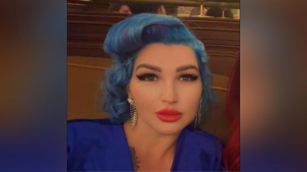 Tania Williams is inside looking into the camera. She has wavy blue hair. She is wearing make-up, including bright red lipstick and black mascara. She is wearing a dark blue dress.