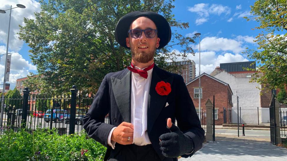 A man dressed in a hat and fingerless glove and bow tie shows the thumbs up sign