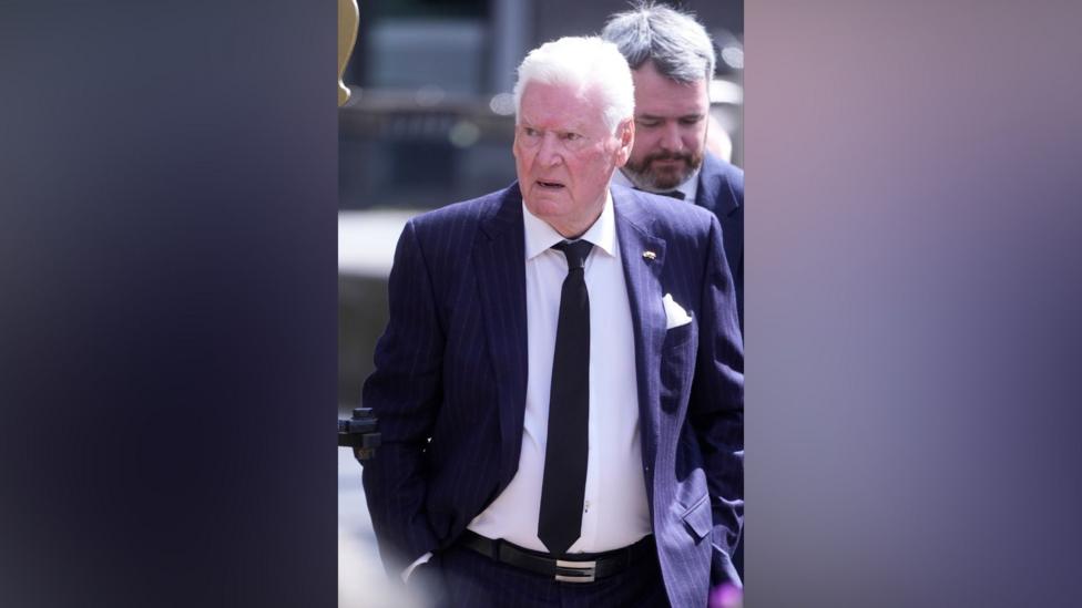 A man with white hair wears a navy suit and black tie