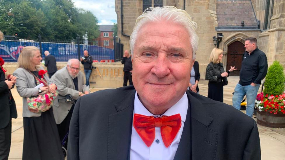A man with grey hair wears a red bow tie and black suit