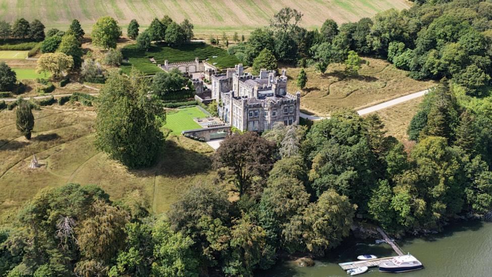 An aerial view of a castle, surrounded by trees and fields. It is overlooking a river.