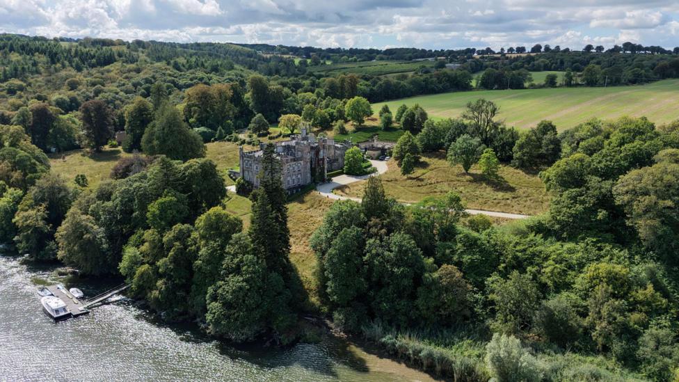An aerial view of a castle, surrounded by trees and fields. It is overlooking a river. There is a boat on the river.