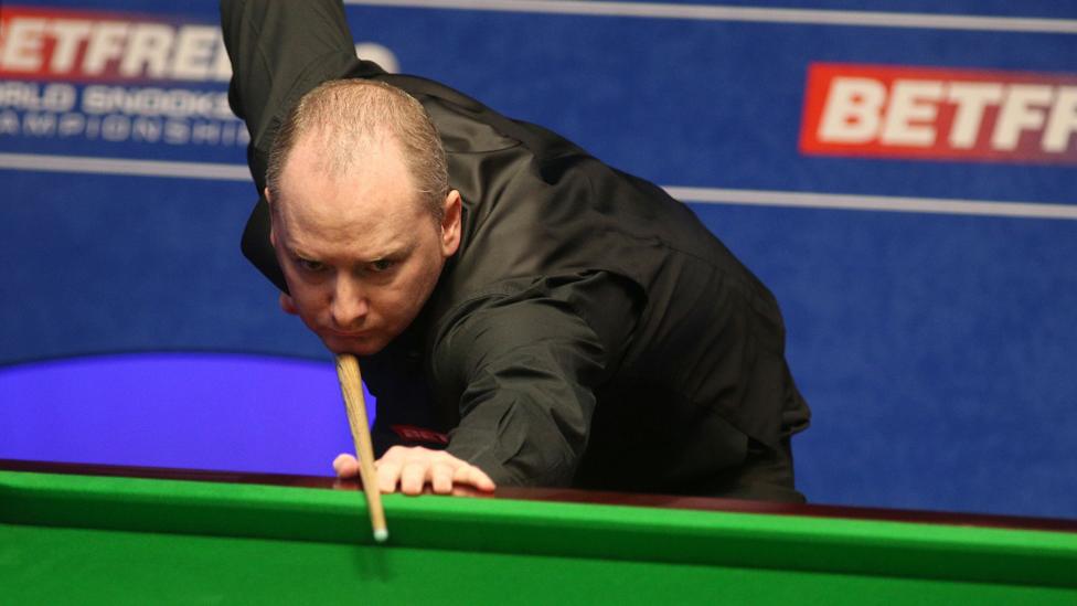 A balding man in a black shirt holds a snooker cue as he prepares to take a shot