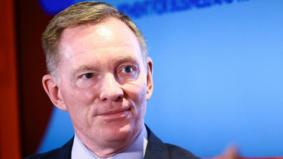 A man with short fair haired wears a black suit and blue shirt. He stands in front of a blue screen.