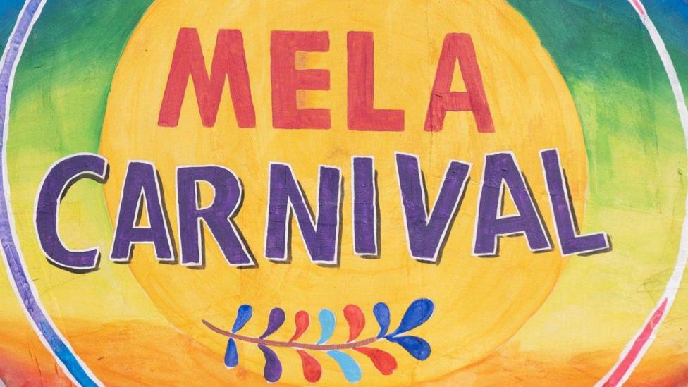 A painted canvas, it has bright colours. The backdrop is a gradient of blue, green into yellow and orange. In large letters it says 'Mela Carnival' in red and purple.