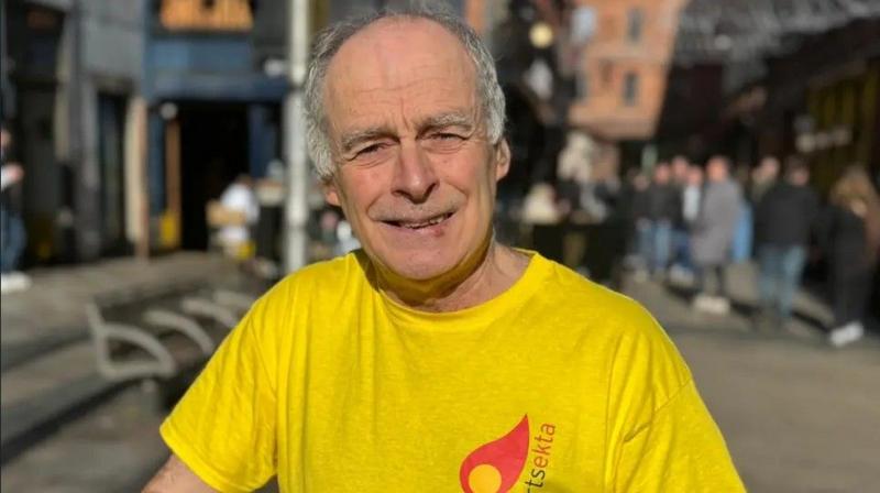 A man, Séamas Ó Labhradha pictured outside in Belfast city centre. He has grey hair and is wearing a yellow top with the arts organisation's logo Arstekta on it in red and black.