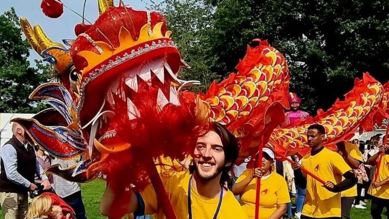 A man, Luke McFarlane at Mela festival pictured with a Chinese dragon. He is smiling, has long brunette hair and is wearing a yellow t-shirt. Behind him are other performers and trees.