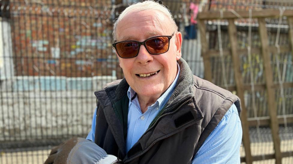 A smiling older man wearing dark sunglasses, a light blue check shirt, and a brown fleece-lined vest stands outdoors on a sunny day. Behind him, a wire fence and brick wall hint at an urban garden setting. His relaxed posture and warm expression convey friendliness and pride, as if he’s enjoying the results of a community project.