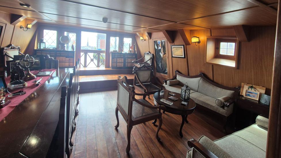 A warmly furnished, wood-paneled interior of is the admiral's quarters on a historic ship, with several chairs and a table.