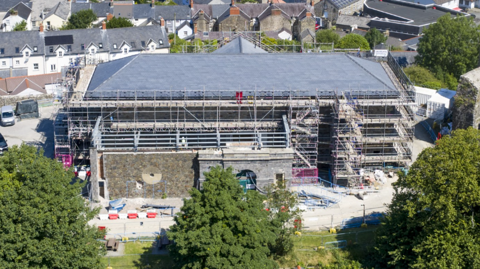 The gaol building undergoing construction across three floors is surrounded by green trees and houses.