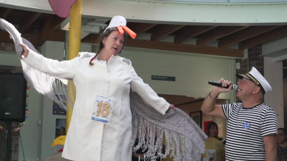 A woman dressed in a white coat with a seagull-shaped hat with shimmery wings is standing on a stage with her arms spread out. A man wearing stripes and a sailor's hat is holding a microphone.