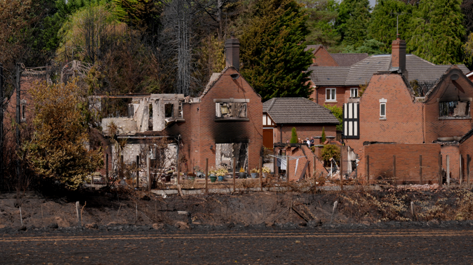 A burnt out house in Stourbridge, which has lost its roof and most of its walls, next to a burnt but not destroyed property. Charred grass is visible in front of the houses.