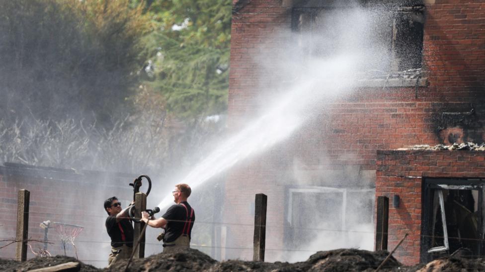 Two male firefighters operate a hose which blasts water at the winder of a house, with burn marks around the window.