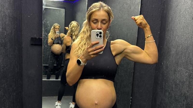 Becki Barratt, who is wearing a sports bra so her baby bump is on display, is posing for a selfie in front of a mirror. She has flexed her left arm and holds the camera phone in the other.