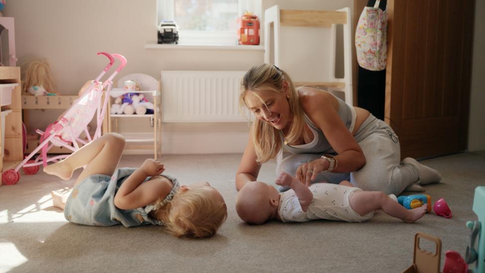 Becki is crouched down over her baby boy in a play room in there house. Her little girl is lying on the floor opposite. There are toys surrounding them.