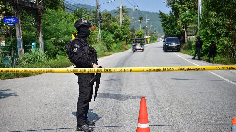 A soldier completely clad in a black uniform and holding a gun stands at a yellow police tape which stretches over a road. A red traffic cone is in the foreground.