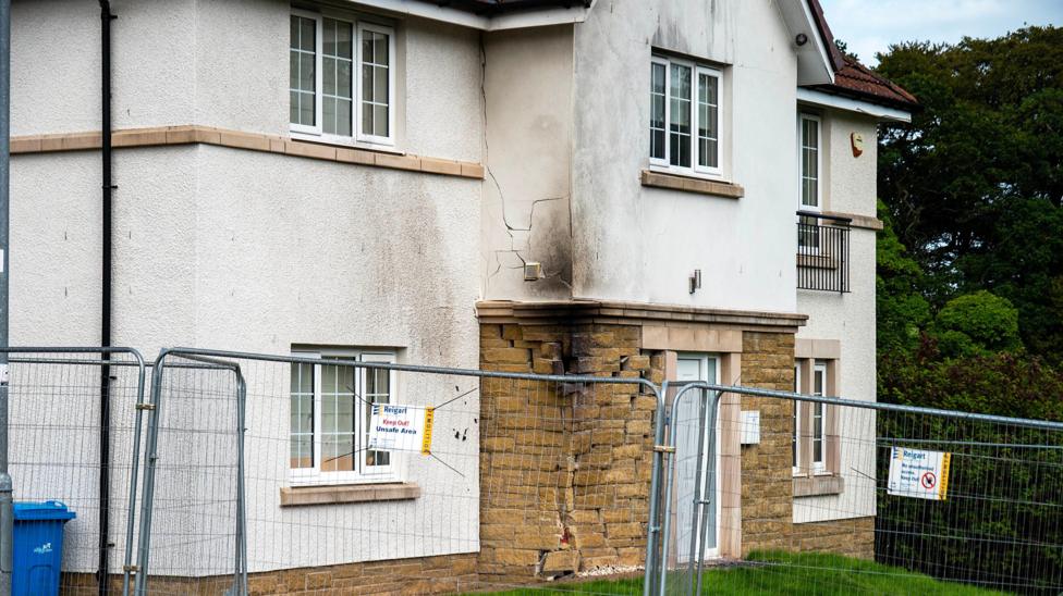 The front of a modern white house which has been damaged by a collision causing structural damage to the stone facing. There are cracks in the white render up the front of the two-storey building and scorch marks all the way up. A high metal fence has been erected in front of the property. 