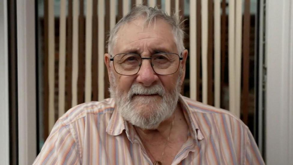 Close-up of Alan, 83, who has closely cropped grey hair and beard and wears large black-rimmed glasses. 