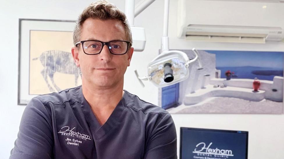 Photo of dentist Jim, a middle-aged man with fair hair and black-rimmed glasses. He's wearing scrubs and standing in a dental treatment room. 