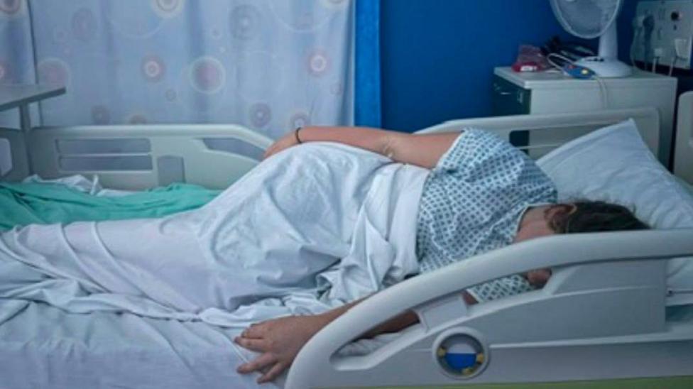 A young woman is lying in a hospital bed wearing a hospital gown and partly covered by a white sheet. We can't see her face as it is hidden by the bed arm. She seems like she may be asleep.