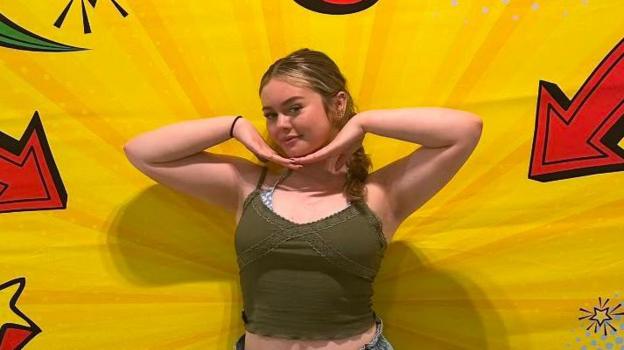 A young woman poses centered against a vibrant, pop-art style backdrop with bright yellow burst patterns and comic-book style red and black arrows or stars pointing inward.