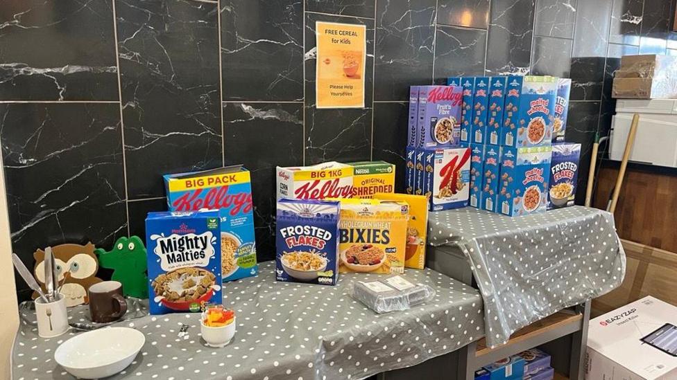 A table with a number of boxes of cereal on, as well as a grey polka dot tablecloth. A sign on the wall reads "Free cereal for kids. Please help yourselves".