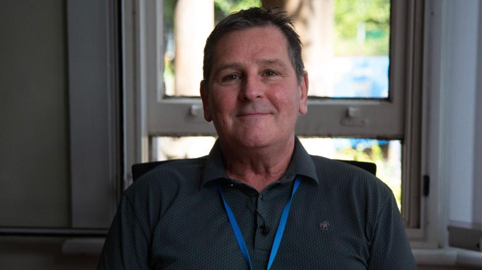 Russell seated indoors in front of a window, wearing a dark collared shirt and blue lanyard. Russell is clean shaven and has short, spikey hair.