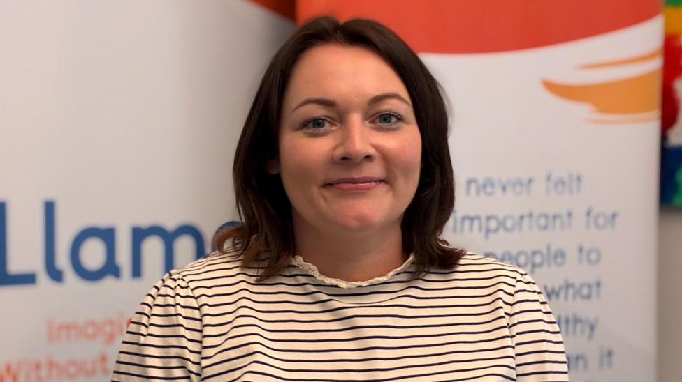 Yvonne photographed indoors in front of promotional display banners, wearing a striped long-sleeved top. Yvonne has shoulder length brown hair and is smiling at the camera.