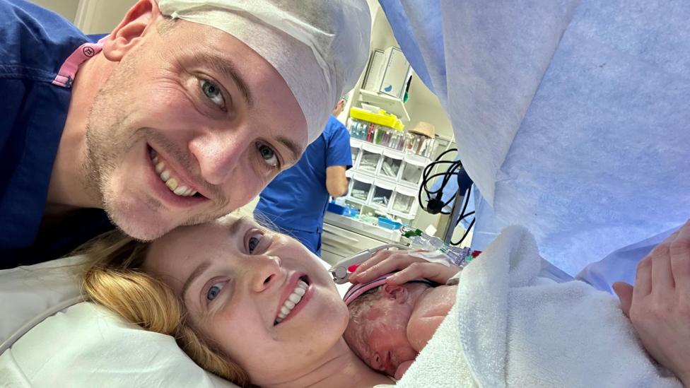 The couple are smiling at the camera in a hospital theatre. Hayley is holding her newborn child.