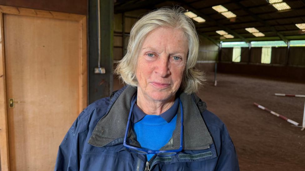 Lady with short hair that's light blonde. She is wearing a blue North Face coat. She has a blue polo shirt and blue jumper. She is standing in a stable.