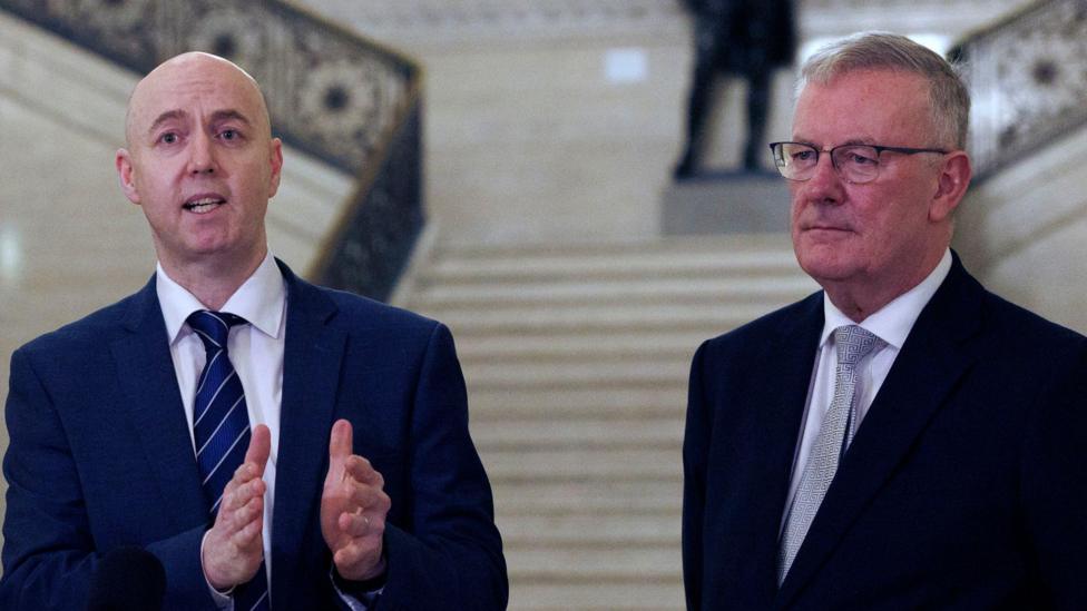 Jon Burrown and Mike Nesbitt. Jon os bald, wearing a white shirt and blue suit. Mike has short grey hair, wearing a white shirt, grey tie and black blazer.