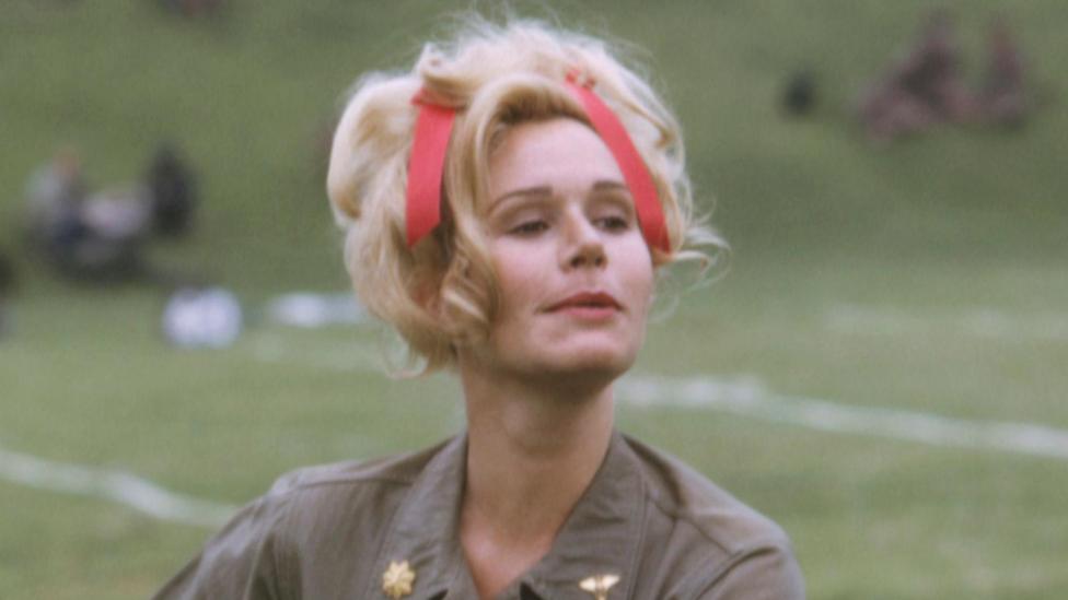 Sally Kellerman performs as "Hot Lips" Houlihan in army khakis with her blonde hair tied up with a red ribbon. She is looking off to the side and appears to be outdoors with grassy scenery behind her