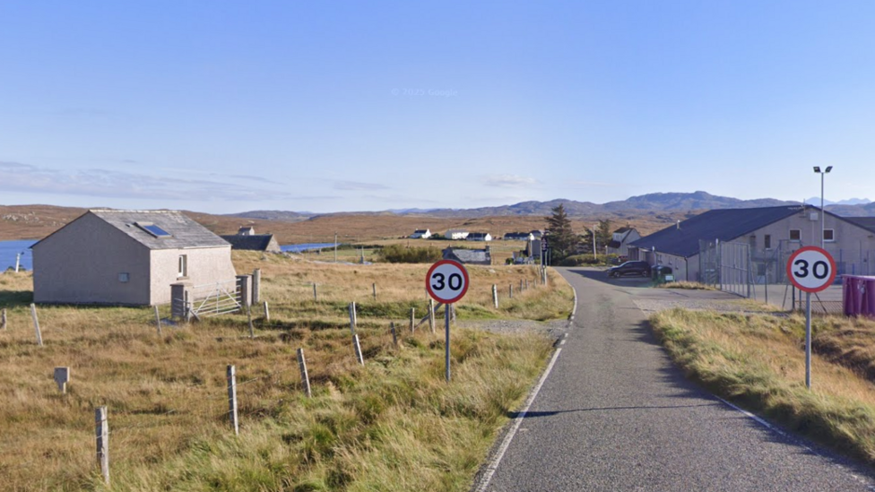 A rural scene showing fields with some small buildings and a body of water in the distance. A road connects the buildings, with two 30mph signs visible in the foreground.