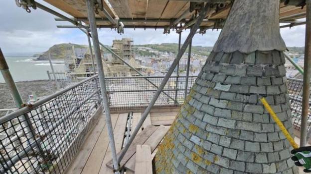 View from a scaffold looking on a slate spire down at the building. A hand with a yellow tape measure is in the lower right corner 