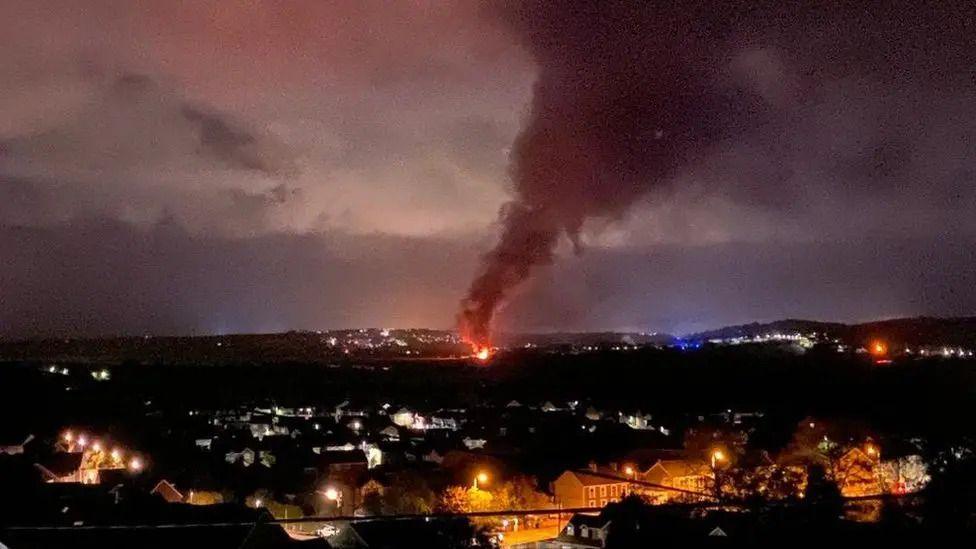 A fire seen in the background with smoke pouring out into the sky. There are houses light by lamp posts in the foreground.