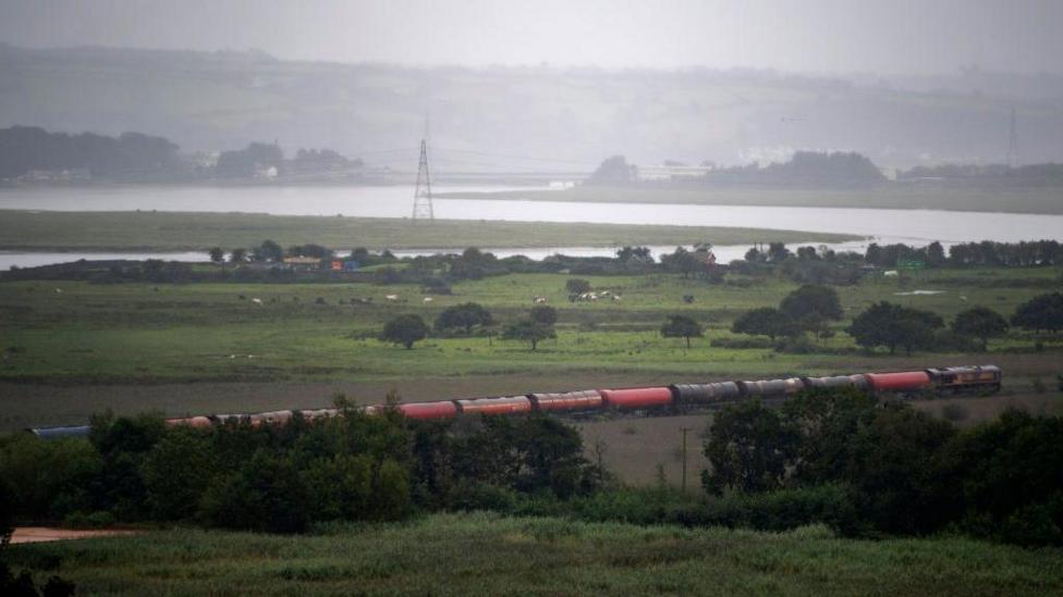 A train travelling through a rural area with a river and marshes seen in the background.