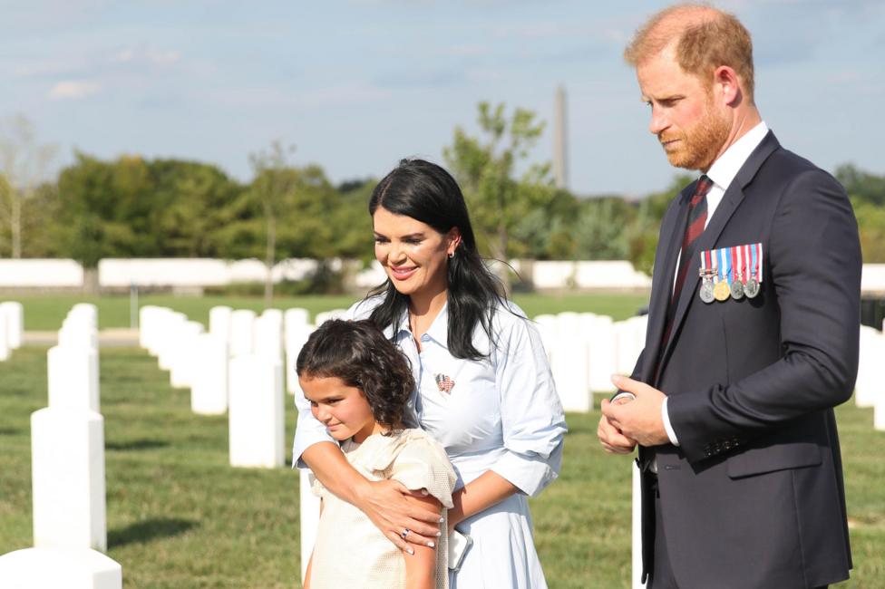 Prince Harry stands in a suit with military medals next to a mother who has one arm around her young daughter. They are standing on a green graveyard honouring military personnel with white gravestones.