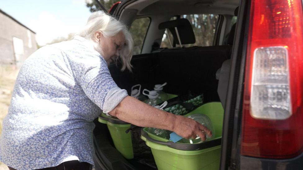 Genista Tanner stands beside the open boot of a small car parked outdoors. Inside the vehicle are several large plastic storage tubs containing water containers and supplies. The person is reaching into one of the tubs. The image illustrates the practical challenges of transporting and storing water after the loss of a private household water supply.