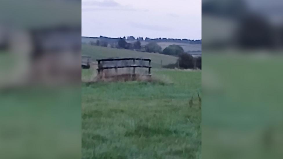A covered well stands in the middle of a green field. Beyond it, rolling farmland, hedgerows and trees stretch across the landscape under an overcast sky. The well is part of the private water supply serving a nearby home.